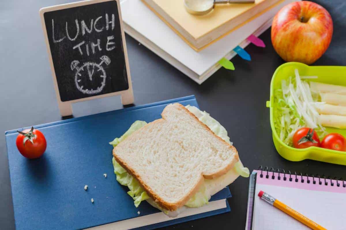 A sandwich on a book, surrounded by a tomato, an apple, a lunchbox with veggies, and a chalkboard reading "LUNCH TIME."
