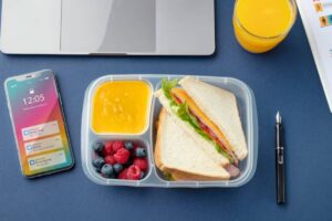A lunchbox with a sandwich, fruit, and orange puree, next to a smartphone showing notifications and a laptop on a blue background.