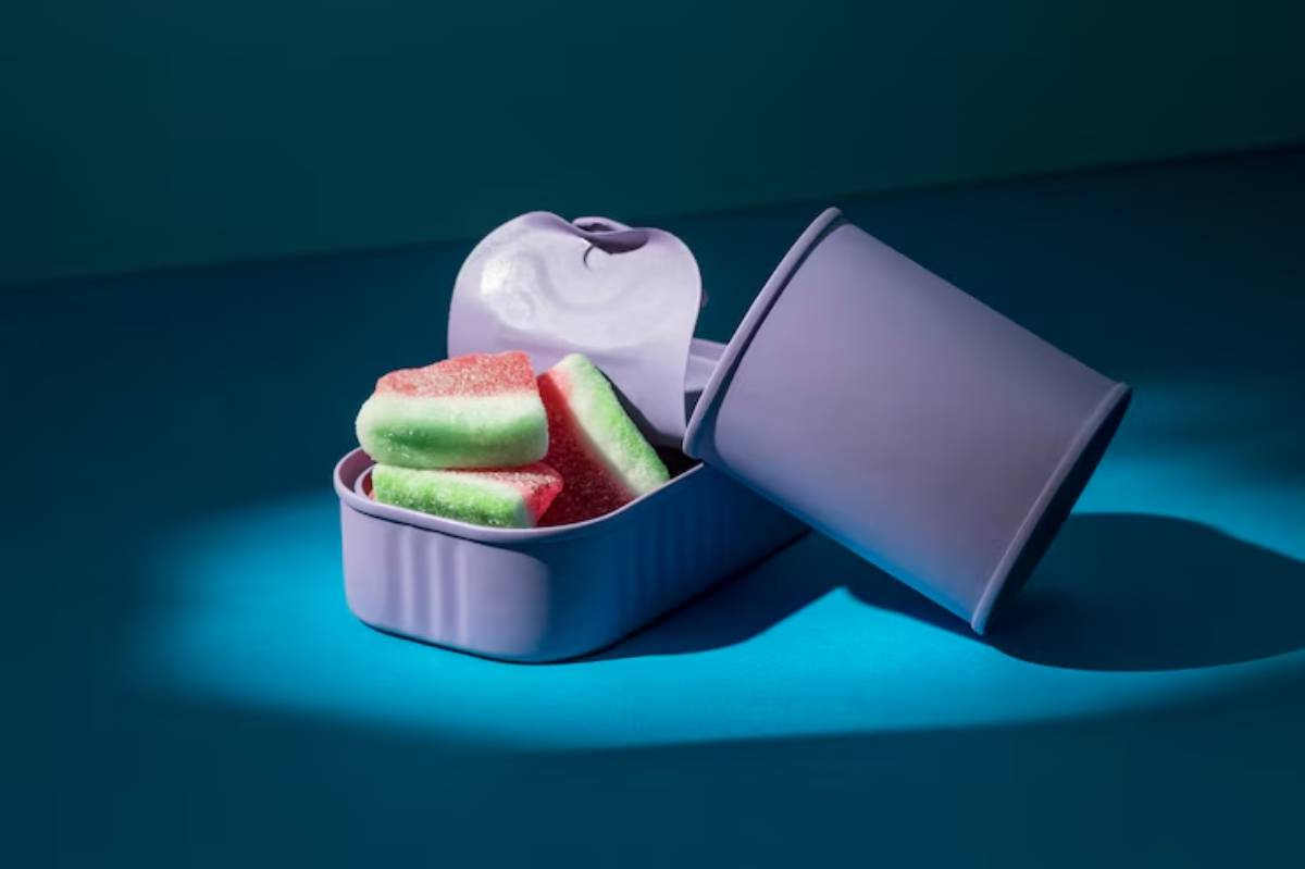 A purple container spills colorful, layered candy pieces resembling watermelon slices, set against a teal background.