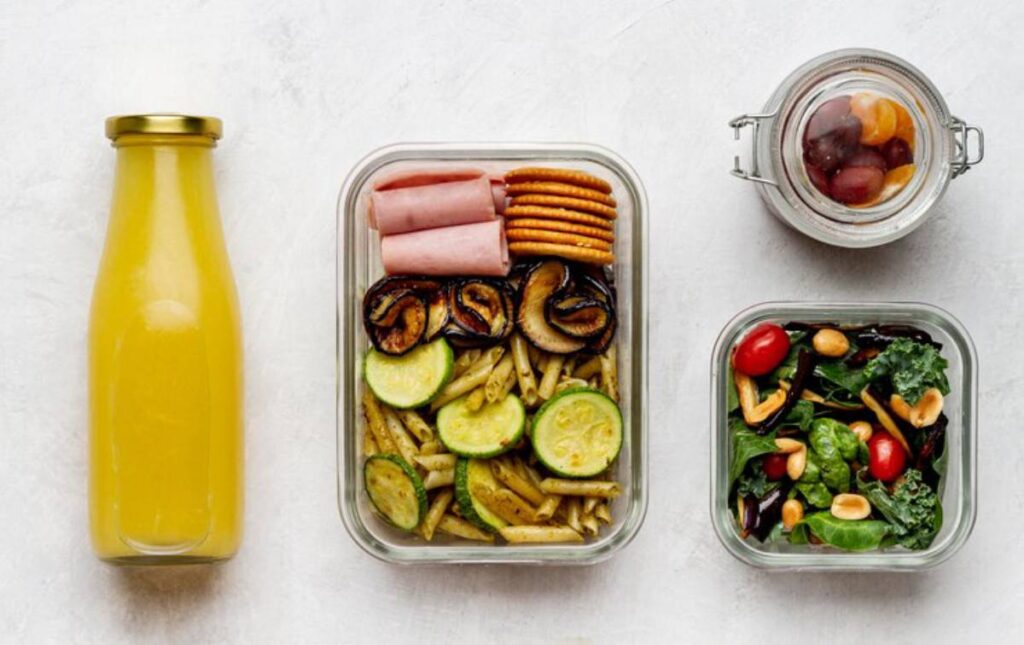 Healthy meal prep featuring a bottle of orange juice, a container with pasta, vegetables, ham, and snacks, plus a salad and fruit jar.