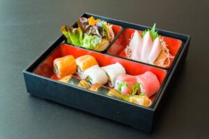 A vibrant sushi platter featuring assorted rolls, sashimi, and a small salad in a divided black bento box against a dark background.