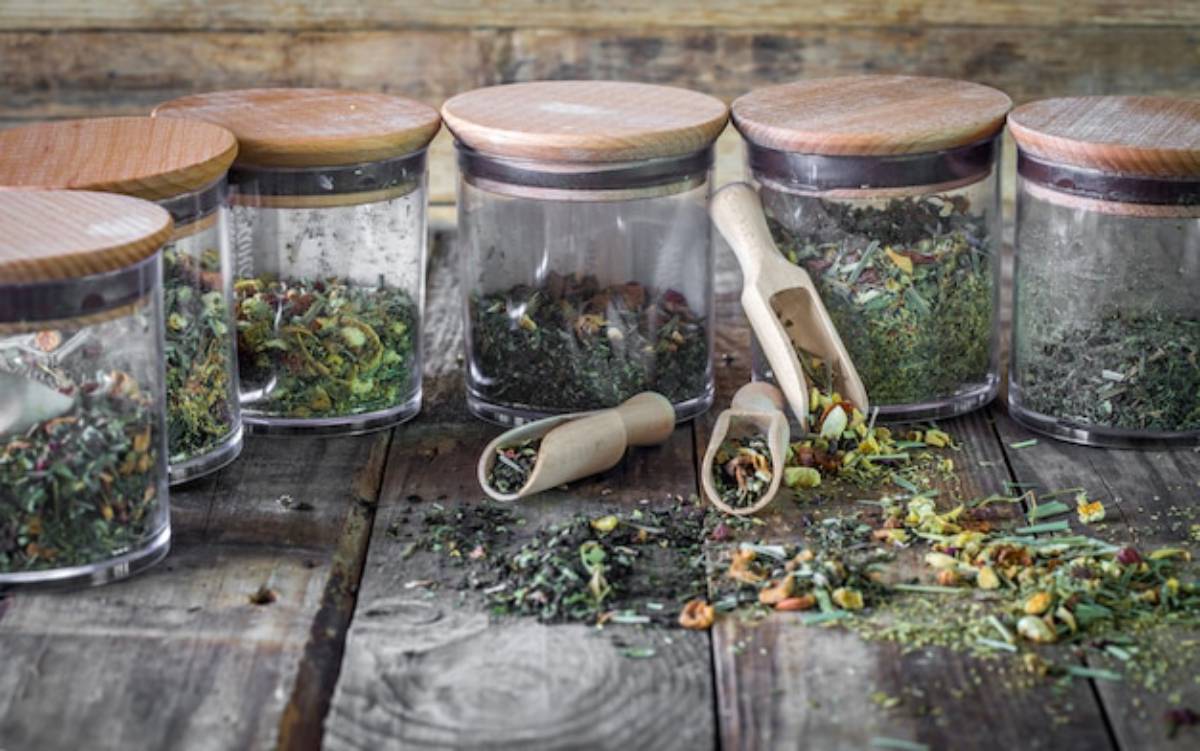 A collection of clear glass jars filled with colorful herbs and spices, accompanied by a wooden scoop on a rustic wooden surface.