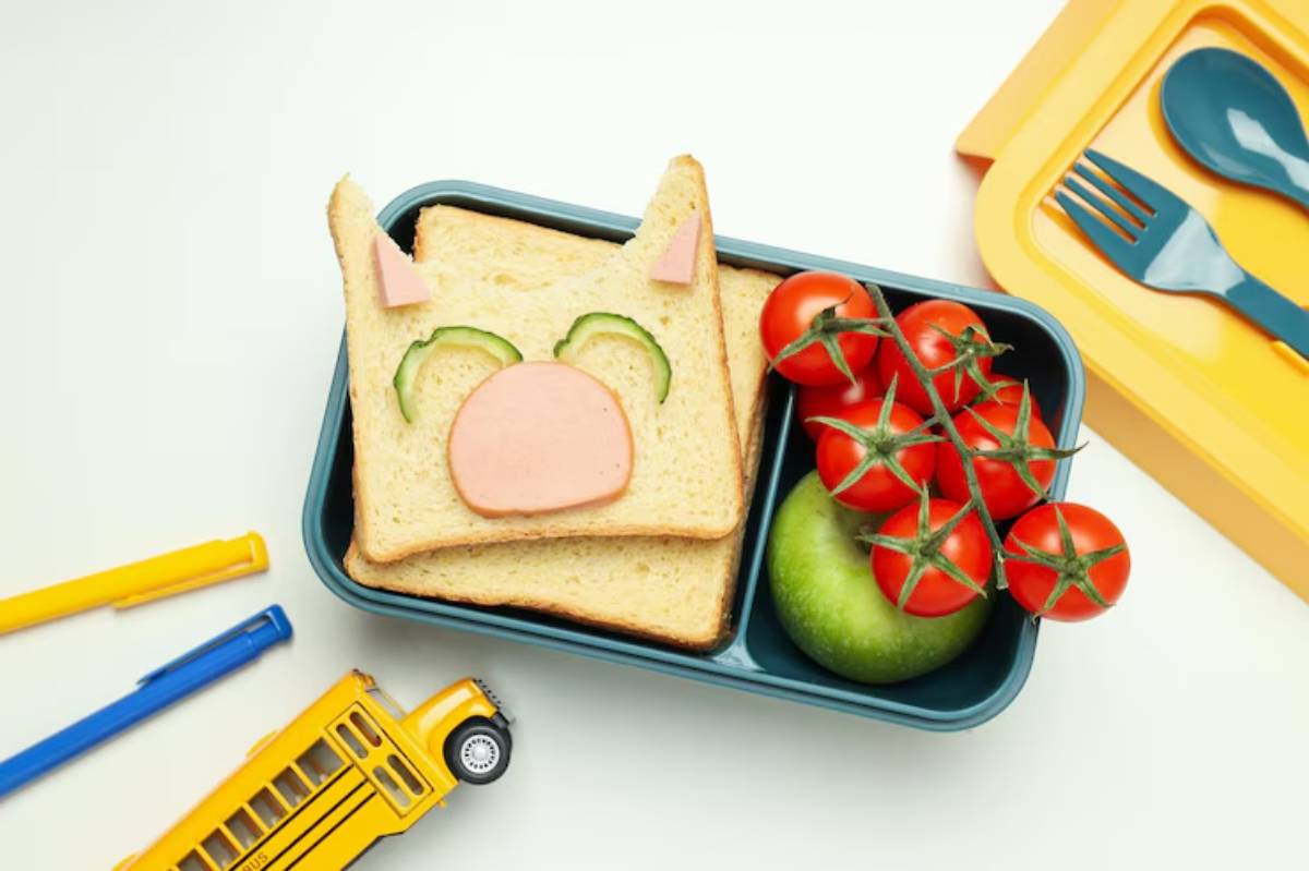 A playful lunchbox with a sandwich shaped like a pig, cherry tomatoes, an apple, and colorful pens next to a toy school bus.
