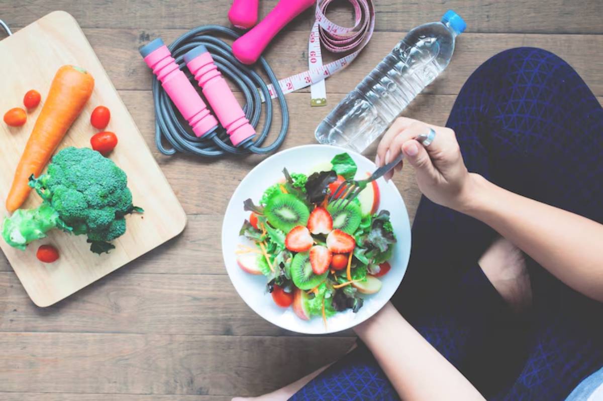 A person enjoying a vibrant salad, surrounded by fresh vegetables, a water bottle, measuring tape, and workout gear.