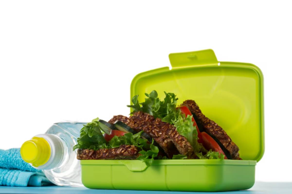 A green lunchbox filled with sesame seed sandwiches, lettuce, and tomatoes, beside a water bottle and a blue towel.