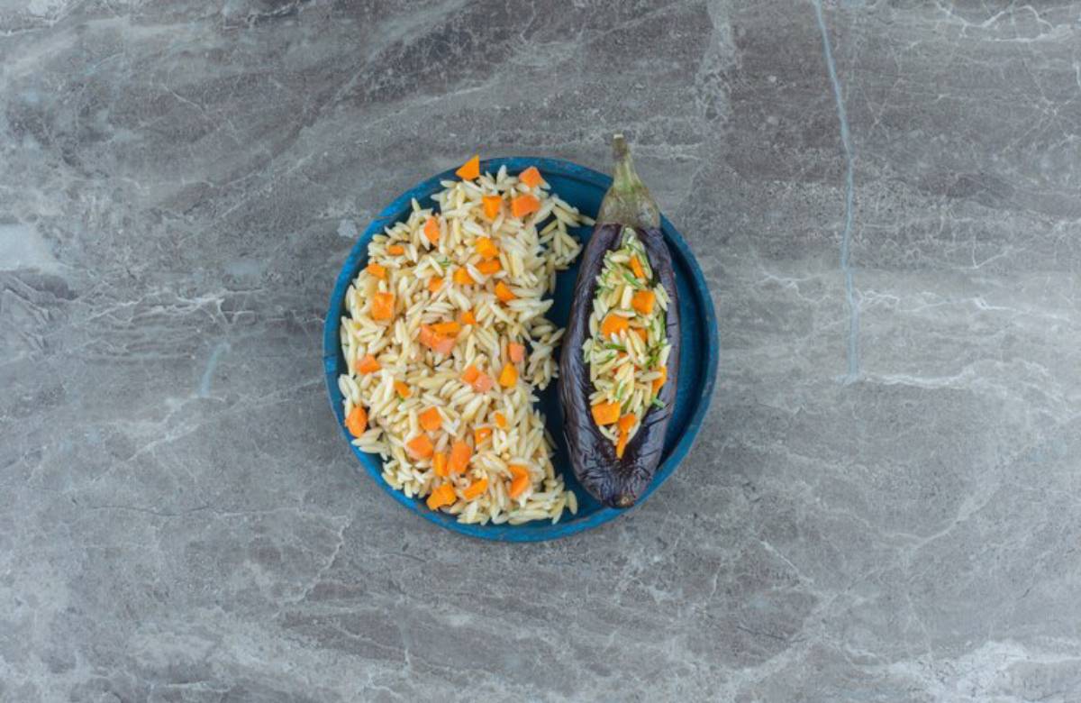 A blue plate with orzo mixed with diced carrots and a stuffed eggplant half, sitting on a gray marble surface.