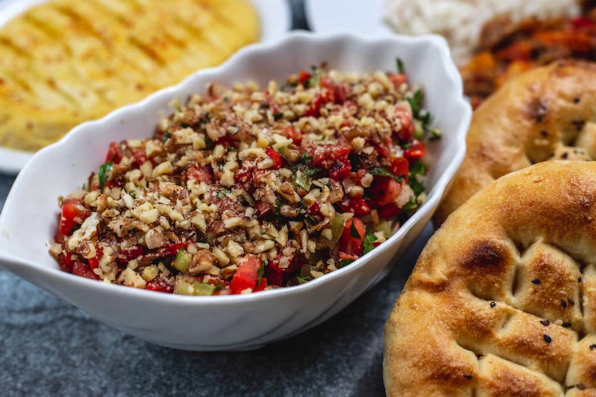 A close-up of a bowl of nut-studded vegetable salad, surrounded by flatbreads and grilled dishes on a dark surface.