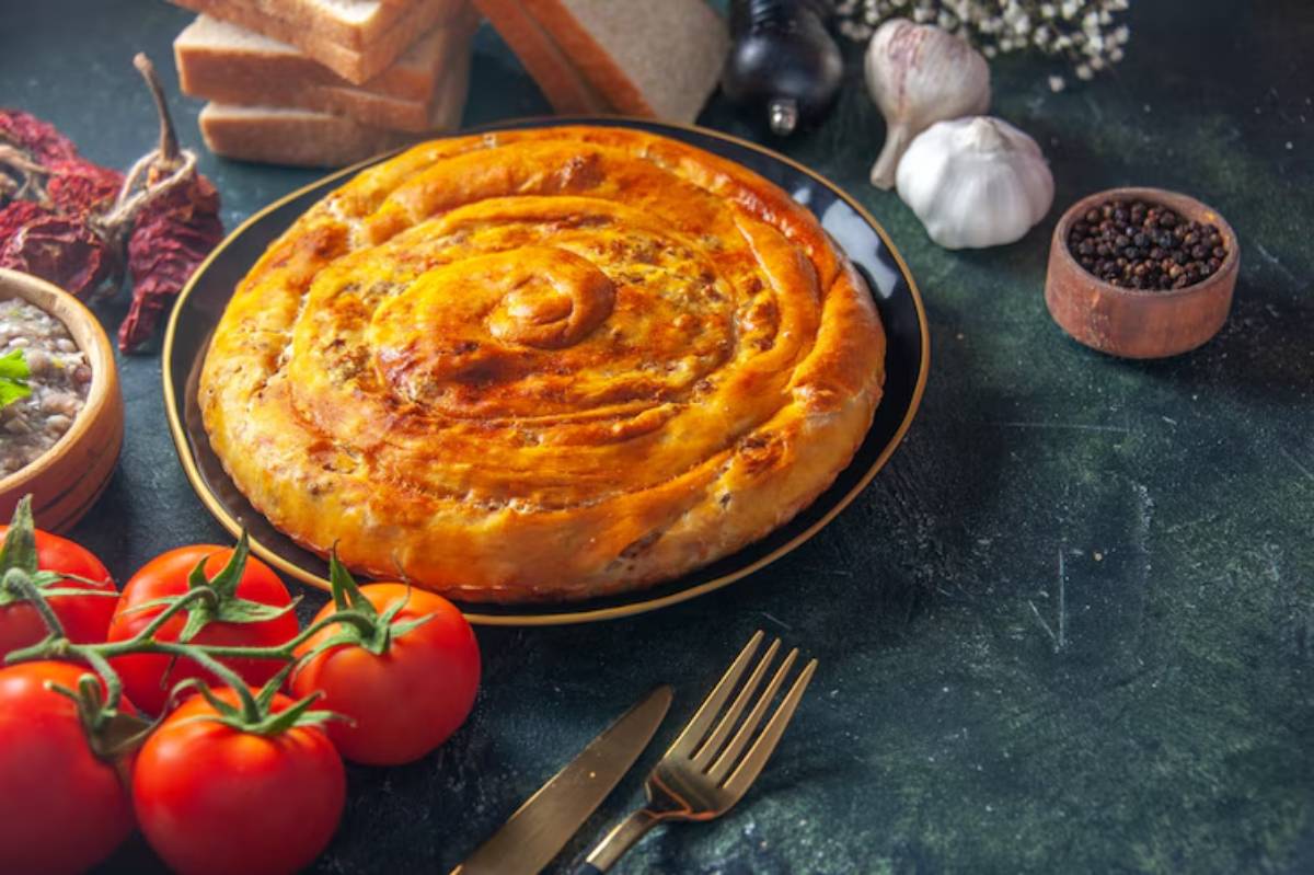 A golden-brown spiral pastry on a black plate, surrounded by fresh tomatoes, garlic, herbs, and spices on a dark countertop.