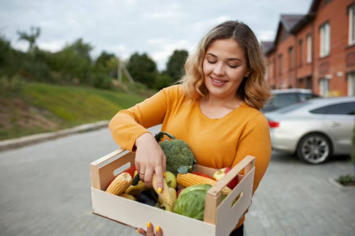 A person in a yellow sweater holds a wooden crate filled with assorted fresh vegetables and fruits, set against a suburban backdrop.