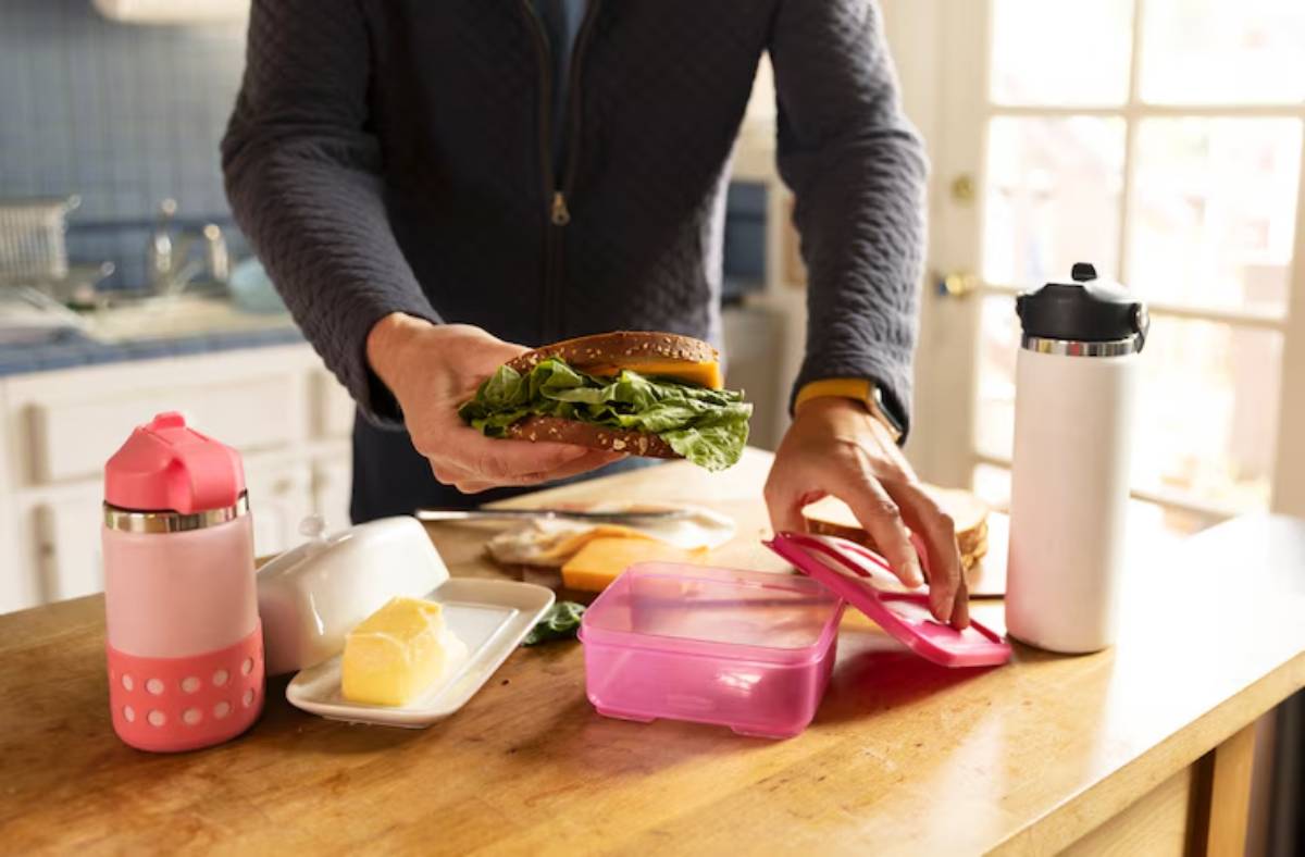 A person prepares a healthy lunch with a sandwich, butter, containers, and a water bottle on a wooden kitchen table.