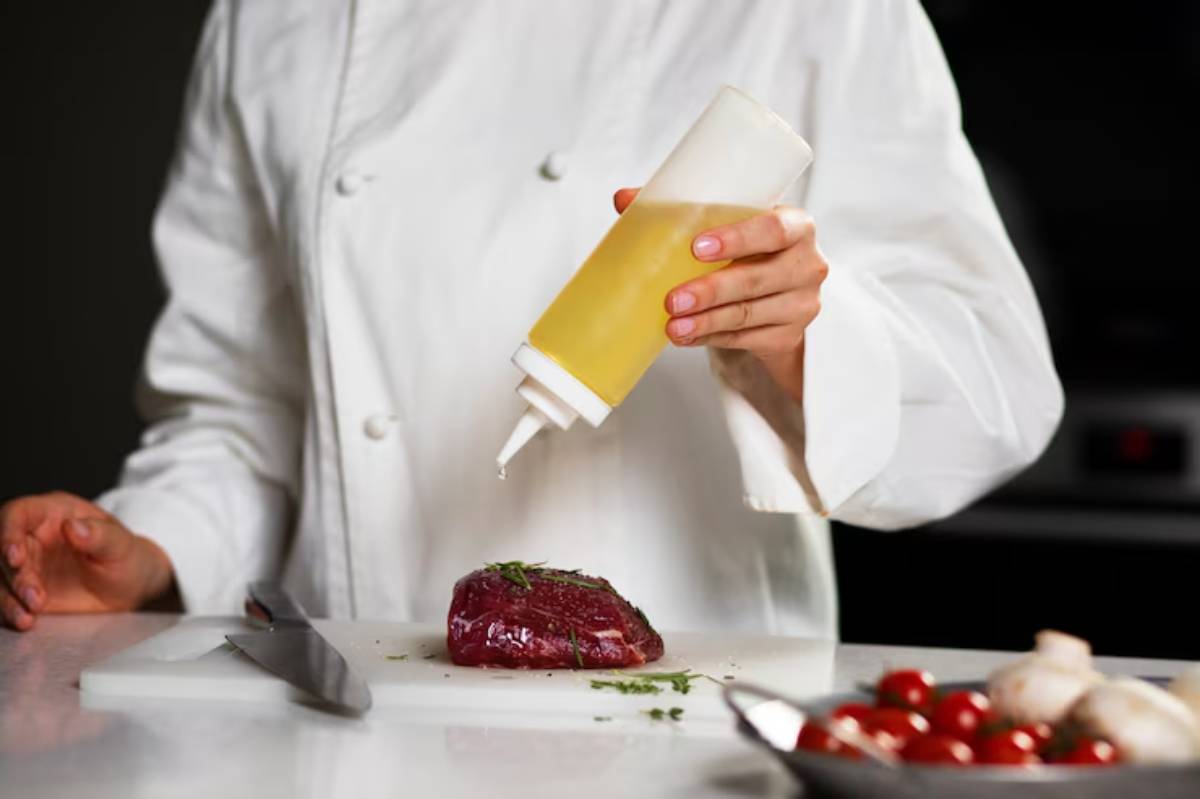 A chef in a white coat drizzles oil from a squeeze bottle onto a seasoned piece of meat on a cutting board, surrounded by tomatoes.