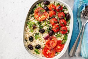 A dish of fluffy rice topped with roasted tomatoes, black olives, and fresh herbs, served with utensils and a blue towel on the side.