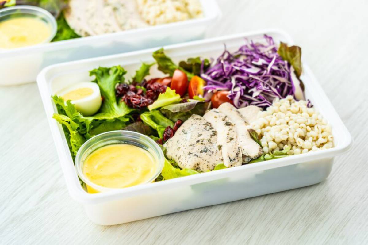 A healthy meal prep box featuring grilled chicken, mixed greens, cherry tomatoes, purple cabbage, grain, and a side of dressing.