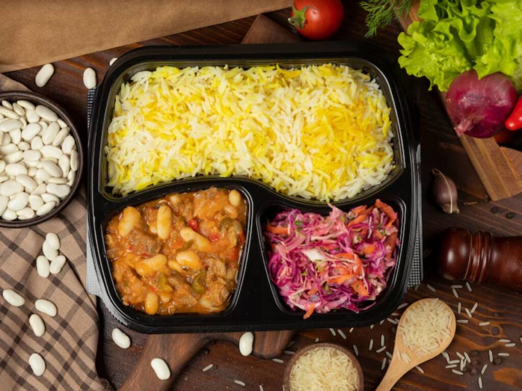 A meal tray features yellow rice, a hearty bean stew, and colorful slaw, surrounded by ingredients like beans, tomatoes, and lettuce.