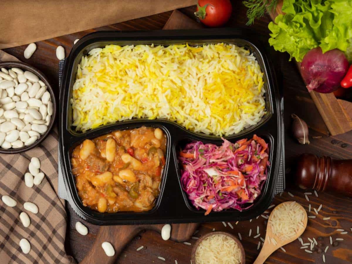 A meal tray features yellow rice, a hearty bean stew, and colorful slaw, surrounded by ingredients like beans, tomatoes, and lettuce.