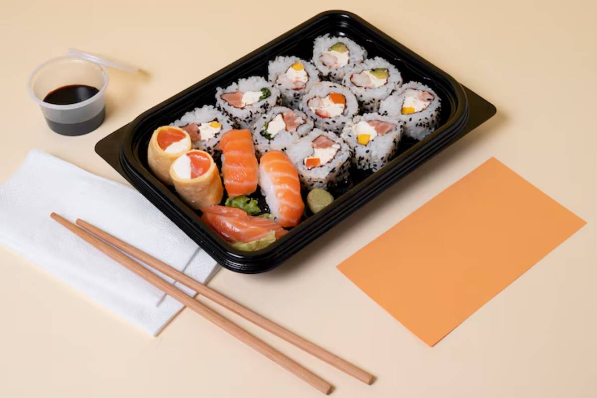 A black sushi container holds various rolls, sashimi, and a small cup of soy sauce, accompanied by chopsticks and a napkin.