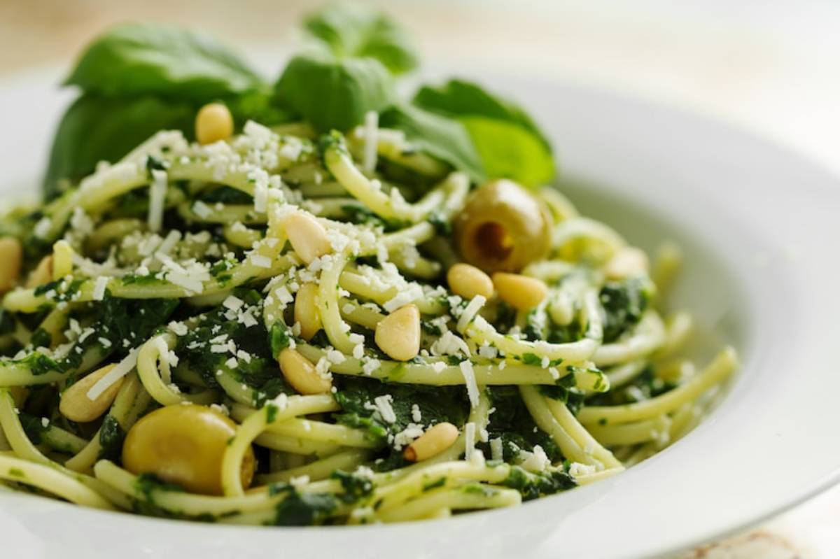 A plate of pasta with green pesto, sprinkled with pine nuts, olives, and grated cheese, garnished with fresh basil leaves.