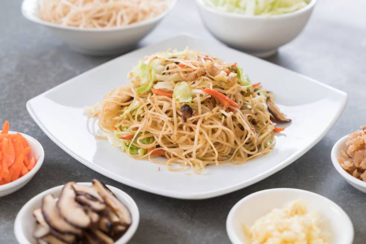 A plate of stir-fried noodles topped with vegetables, surrounded by small bowls of carrots, mushrooms, and other toppings.