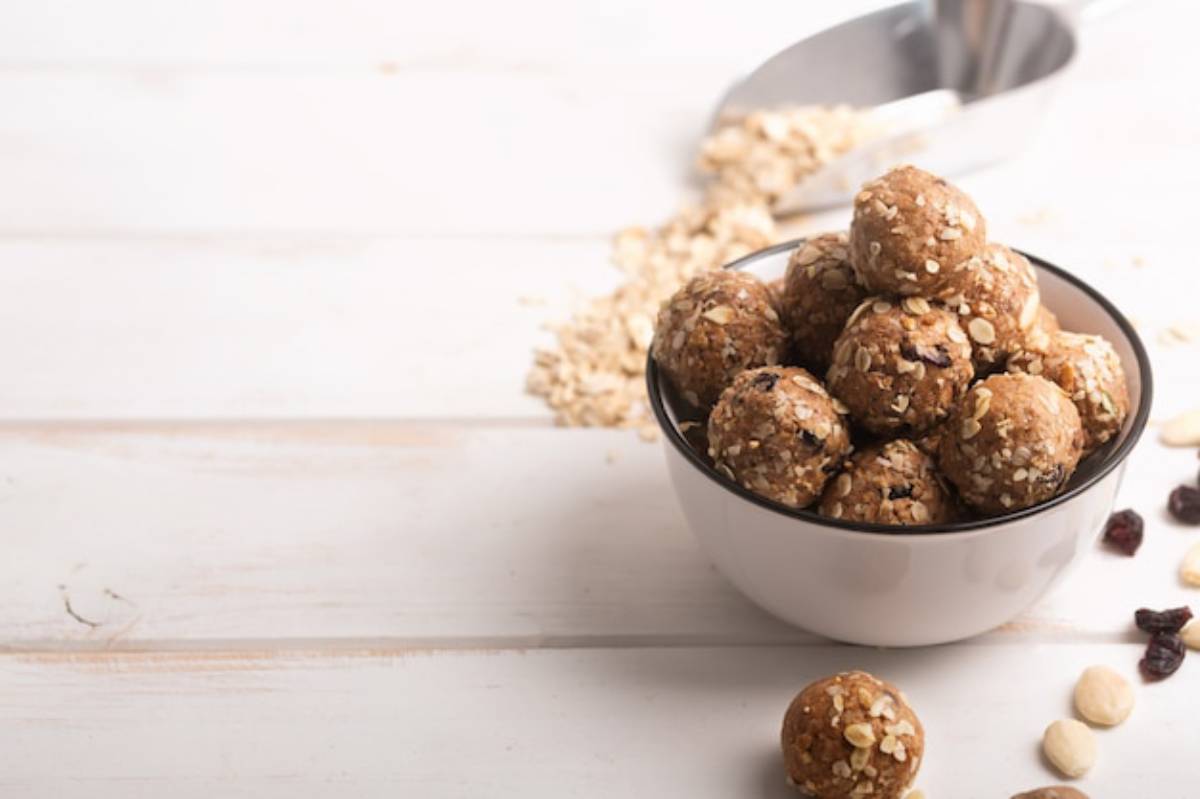 A bowl filled with energy balls made of oats, nuts, and dried fruits, surrounded by scattered oats and ingredients on a white wooden surface.