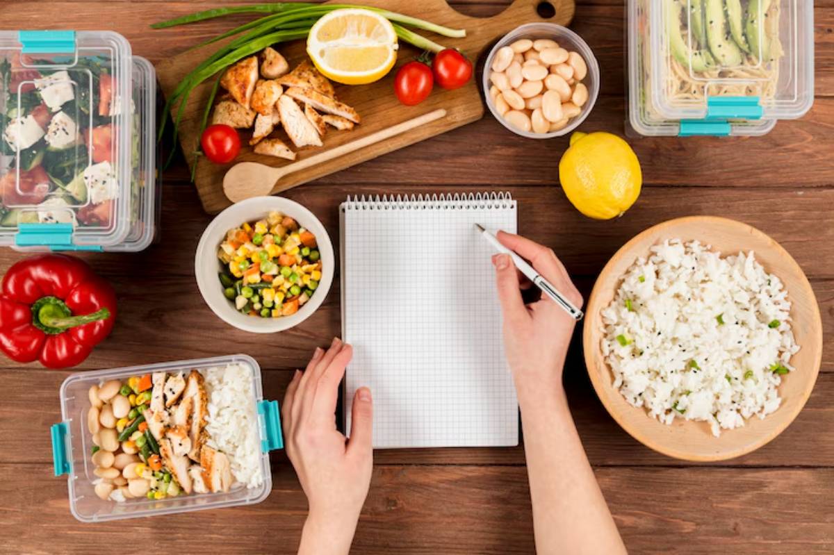 A wooden table displays various food items: containers of salad, grilled chicken, rice, veggies, fresh lemon, and a notepad for meal planning.