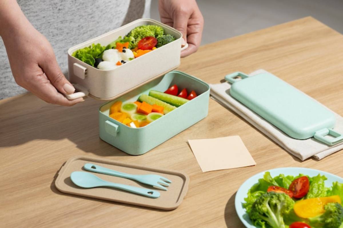 A person holds two cute bento boxes filled with colorful vegetables on a wooden table, alongside utensils and a small note.