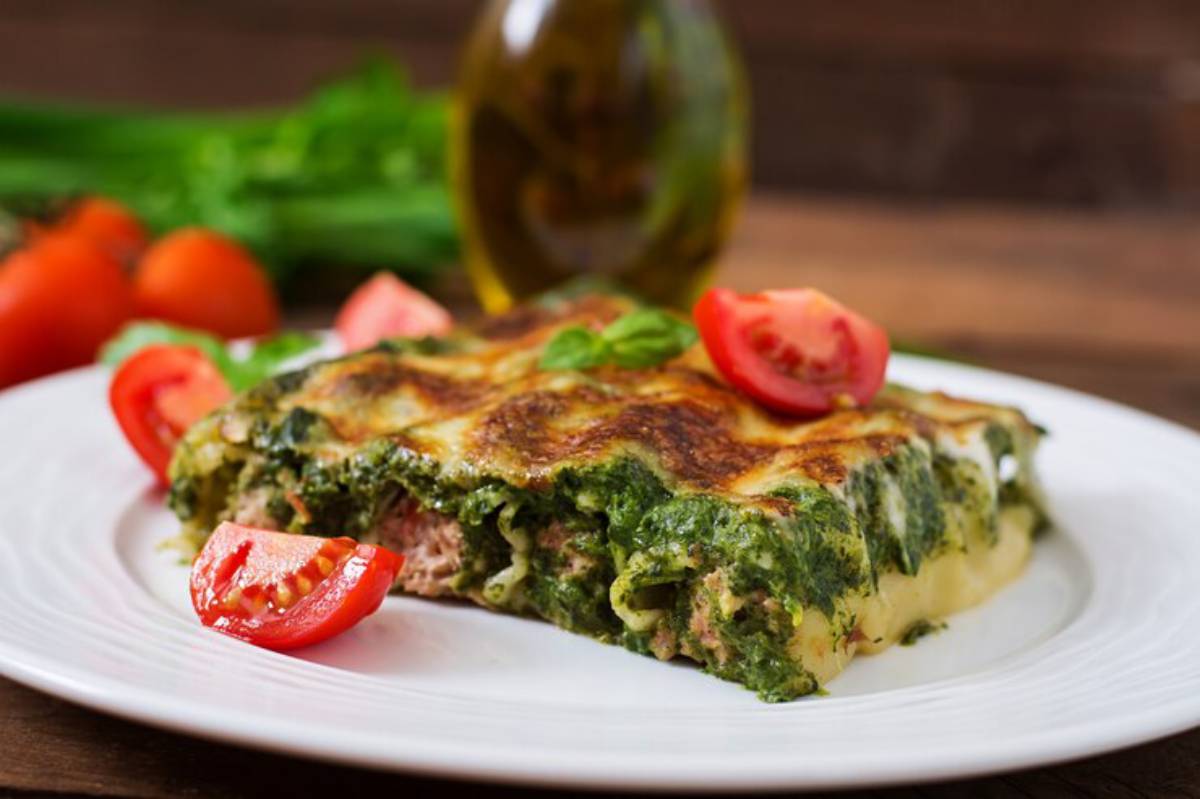 A slice of spinach lasagna on a white plate, garnished with fresh tomato slices and basil, with olive oil in the background.