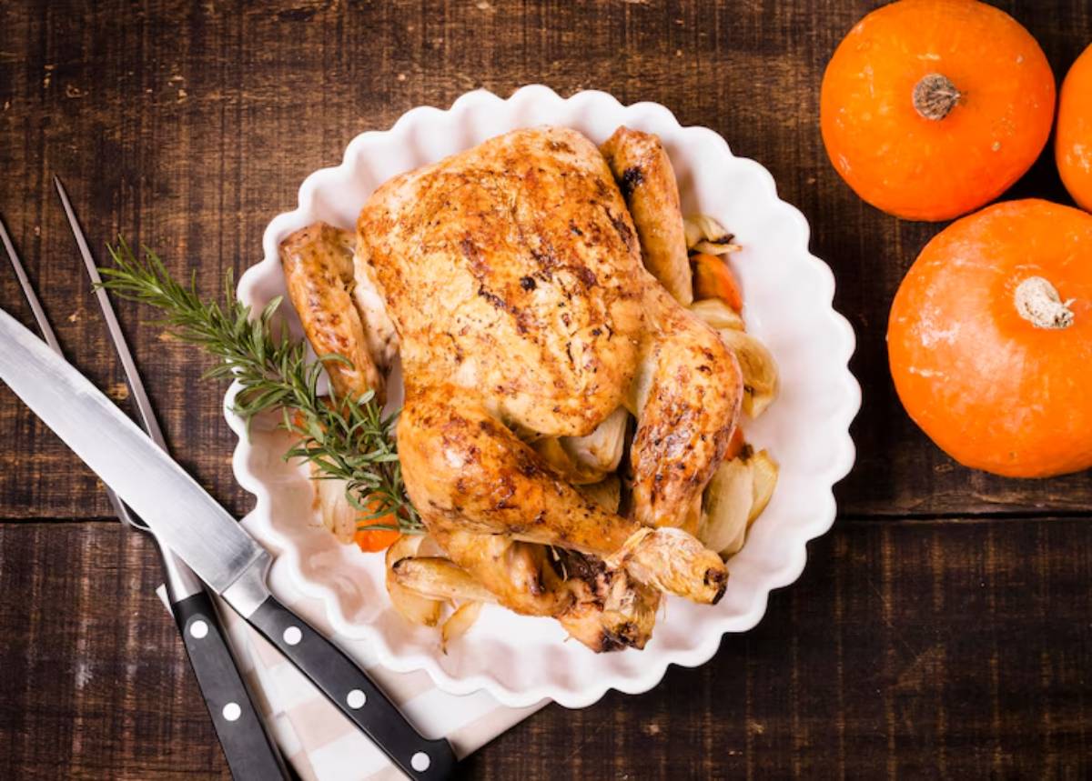 A golden-brown roasted chicken garnished with rosemary on a white platter, accompanied by orange pumpkins and knives on a rustic table.