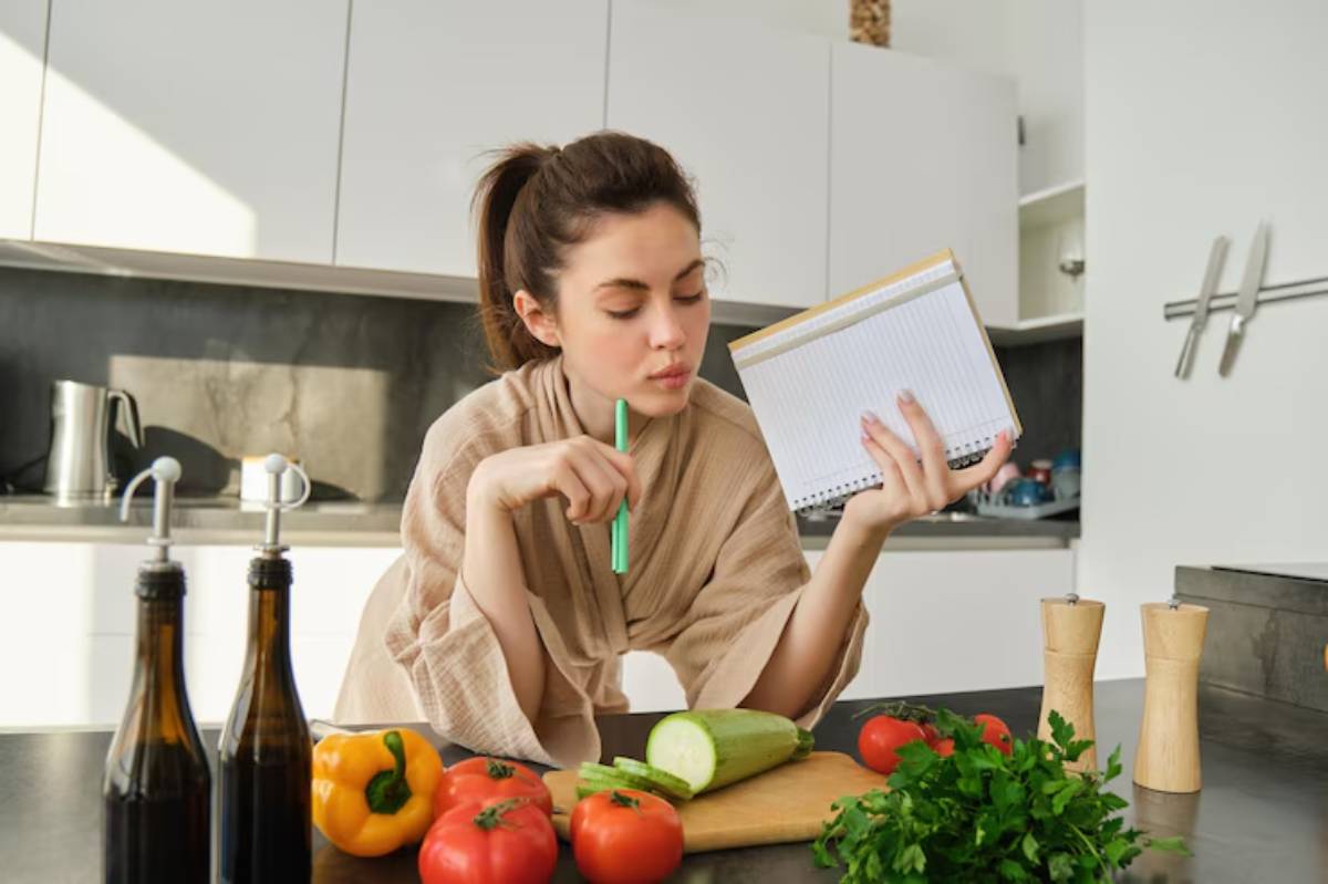 A woman in a kitchen, holding a notebook and pen, surrounded by fresh vegetables like tomatoes, cucumbers, and peppers on a cutting board.
