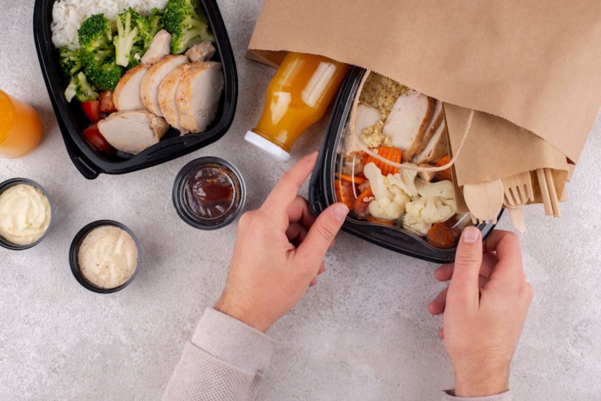 A person is unpacking a healthy meal, featuring chicken, vegetables, rice, and sauces, alongside a juice bottle and utensils in a bag.
