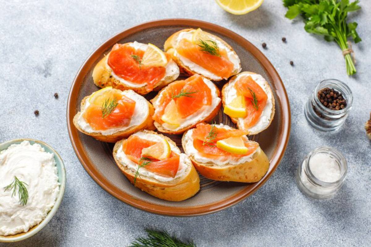 A plate of toasted bread topped with cream cheese, smoked salmon, lemon slices, and fresh dill, surrounded by spices and herbs.