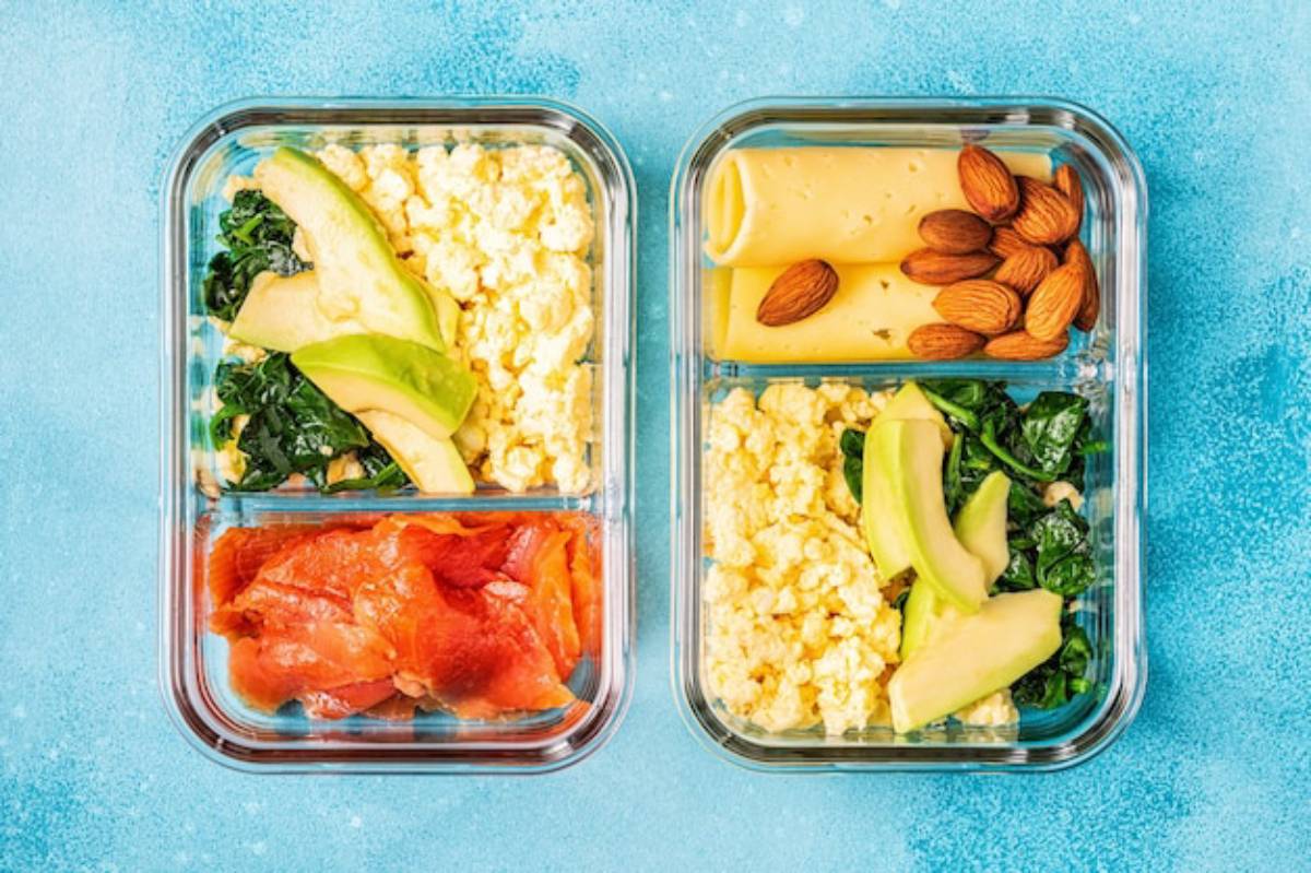 Two divided glass meal prep containers filled with scrambled eggs, avocado, kale, smoked salmon, cheese, and almonds on a blue background.