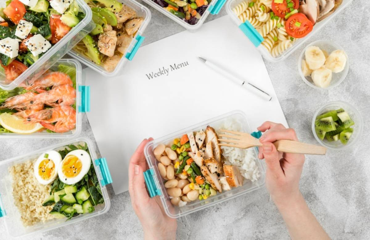 A hand holds a fork above a meal prep container filled with chicken, rice, and vegetables, surrounded by colorful dishes and a weekly menu.