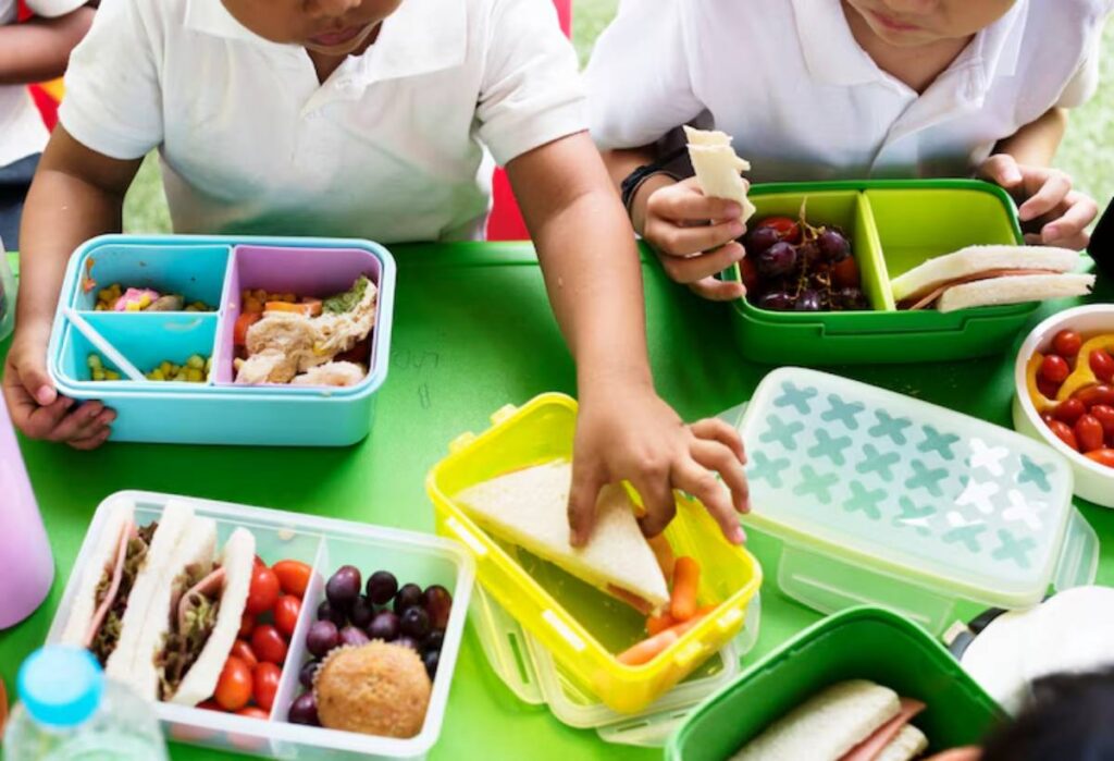 Two children with lunchboxes on a green table, sharing sandwiches, fruits, and veggies during a vibrant outdoor meal.