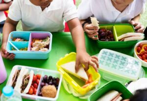 Two children with lunchboxes on a green table, sharing sandwiches, fruits, and veggies during a vibrant outdoor meal.