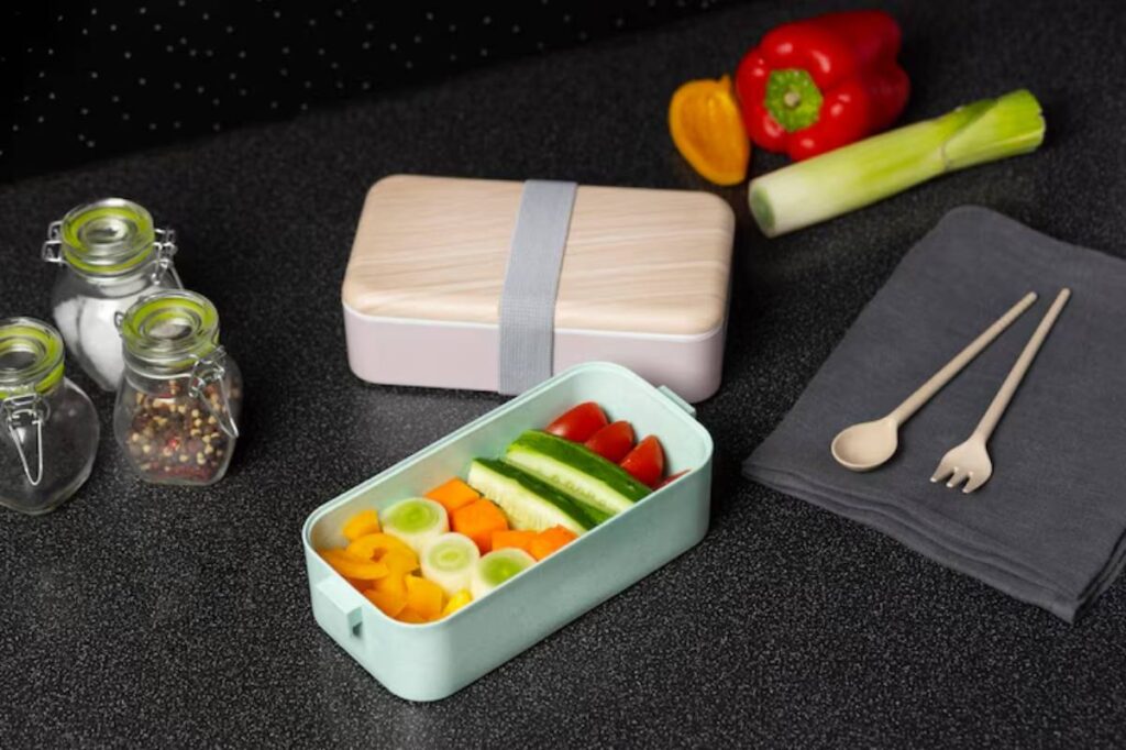 A pastel lunchbox filled with colorful veggies sits on a dark countertop, accompanied by seasonings, utensils, and a napkin.