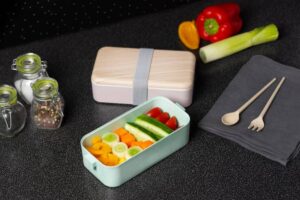A pastel lunchbox filled with colorful veggies sits on a dark countertop, accompanied by seasonings, utensils, and a napkin.