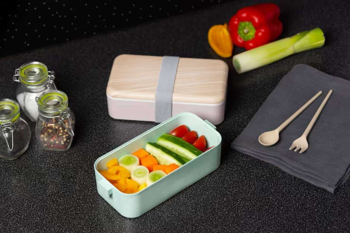 A pastel lunchbox filled with colorful veggies sits on a dark countertop, accompanied by seasonings, utensils, and a napkin.