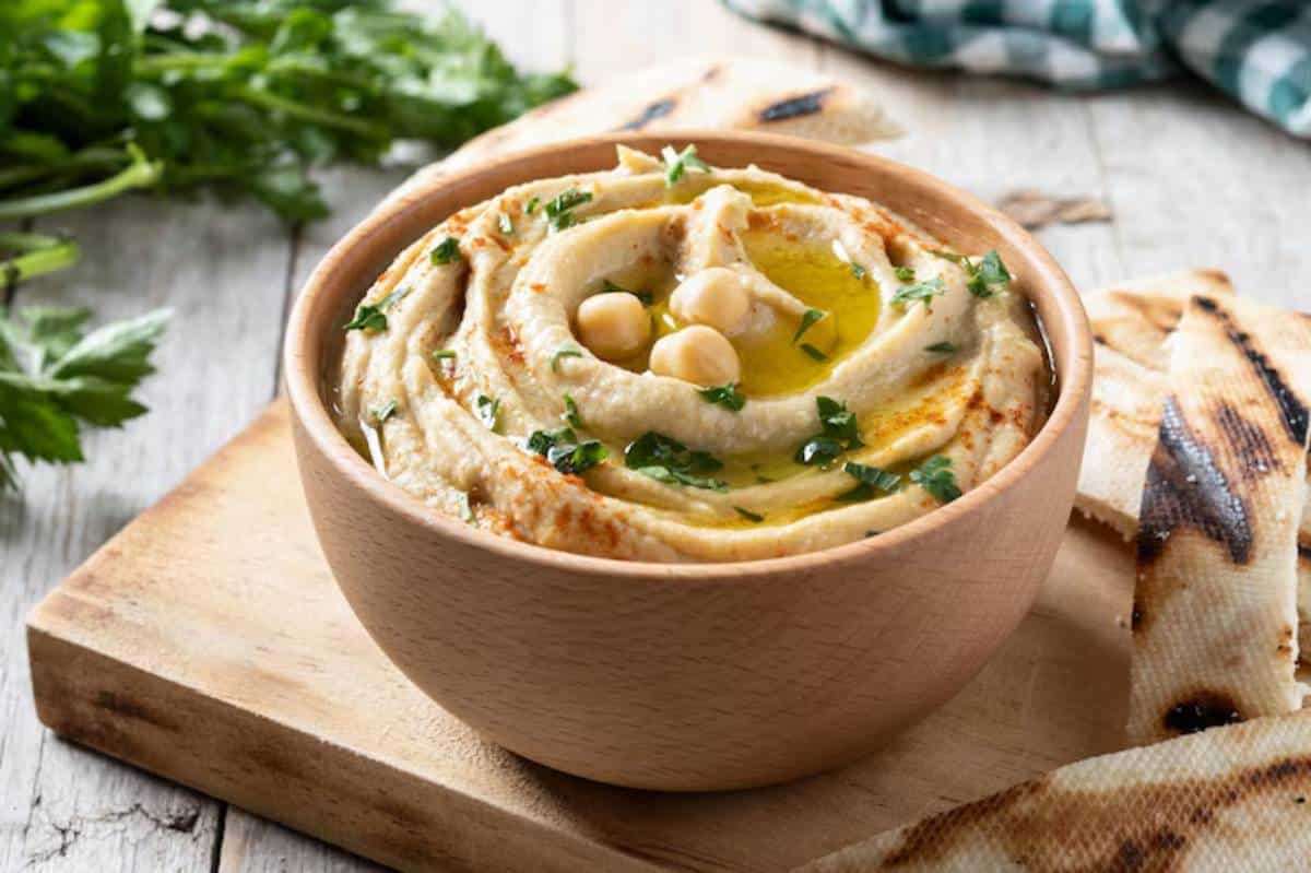 A wooden bowl filled with creamy hummus, topped with olive oil, chickpeas, and chopped herbs, accompanied by grilled flatbread.