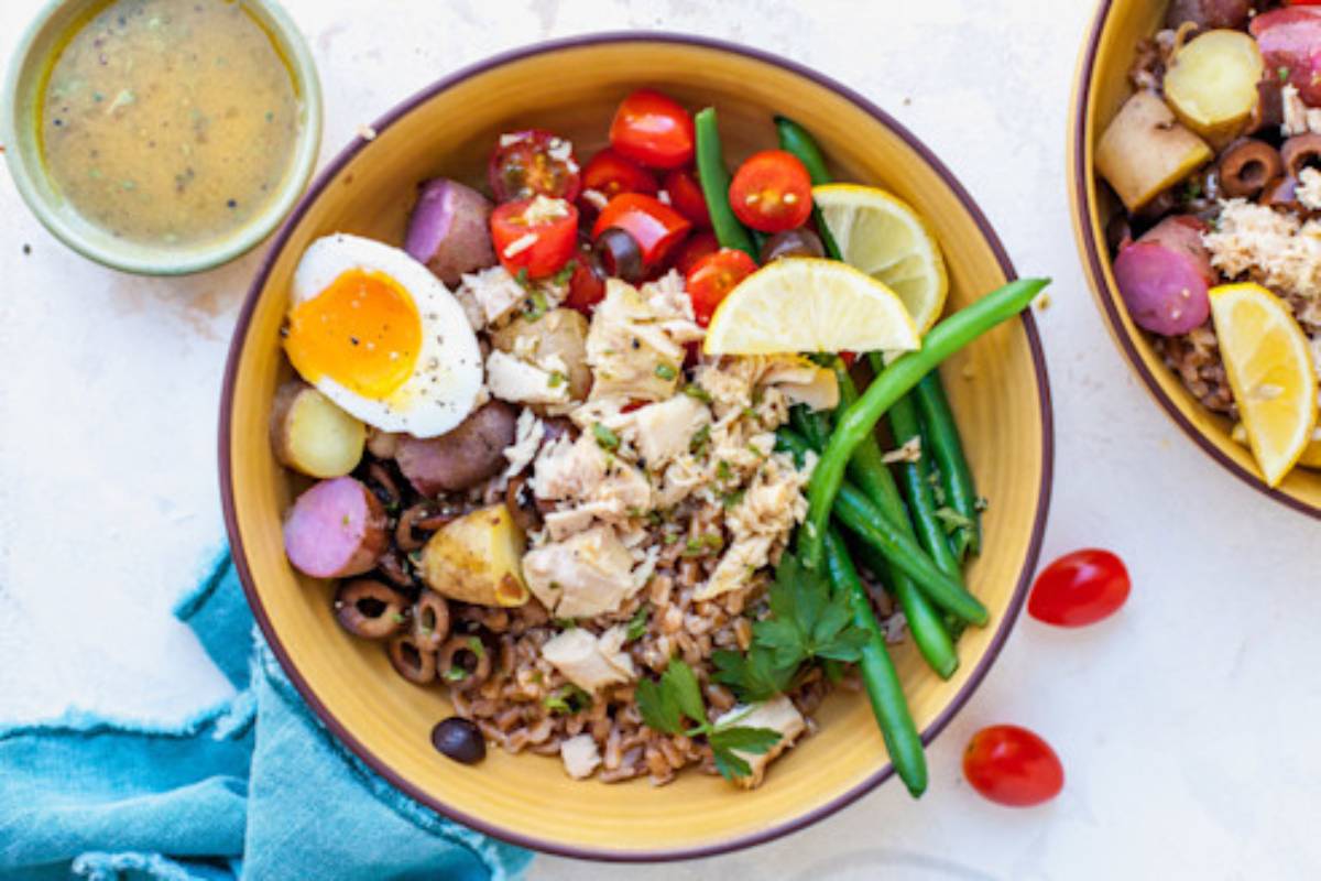 A colorful bowl filled with brown rice, shredded chicken, cherry tomatoes, greens, egg, and lemon slices, with a dressing on the side.