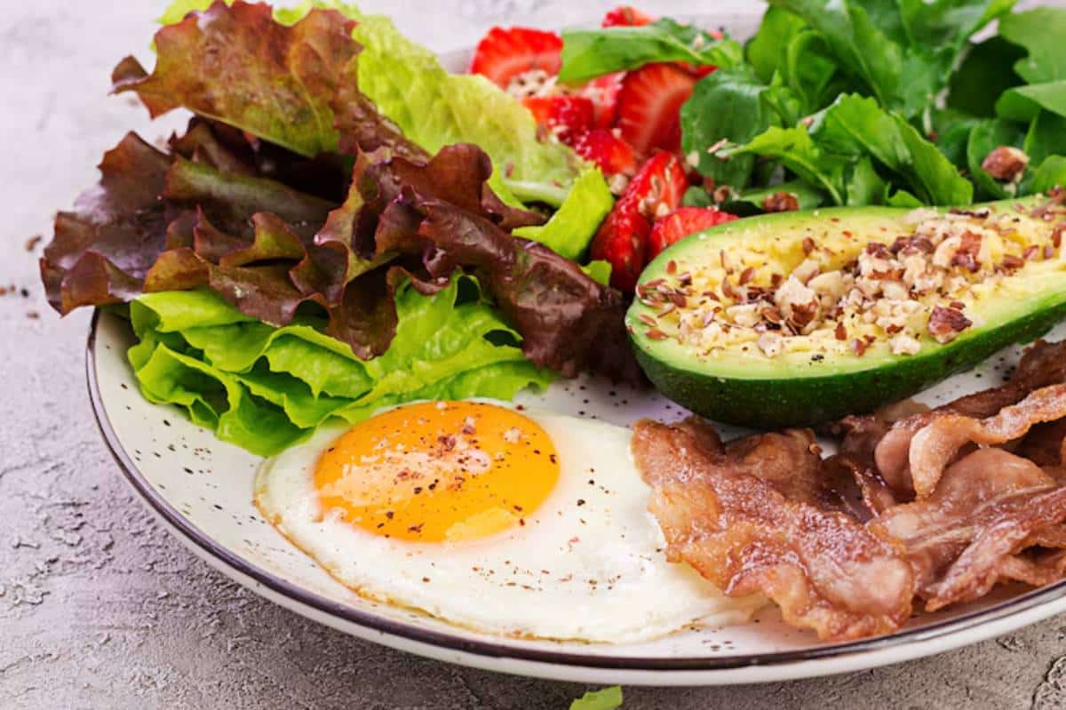A colorful breakfast plate featuring fried egg, crispy bacon, fresh greens, sliced strawberries, and avocado topped with nuts.