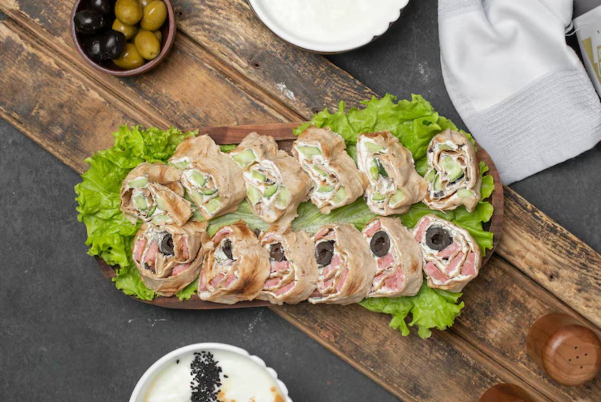 A wooden platter piled with rolled flatbreads filled with meats, olives, and veggies, garnished with fresh lettuce. Small bowls of olives and yogurt are nearby.