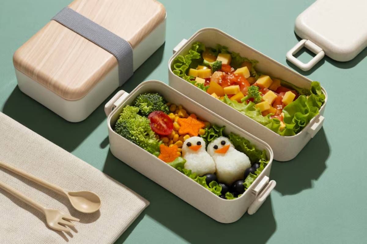 A playful bento box filled with colorful veggies and cute rice penguins garnished with lettuce, arranged creatively on a green surface.
