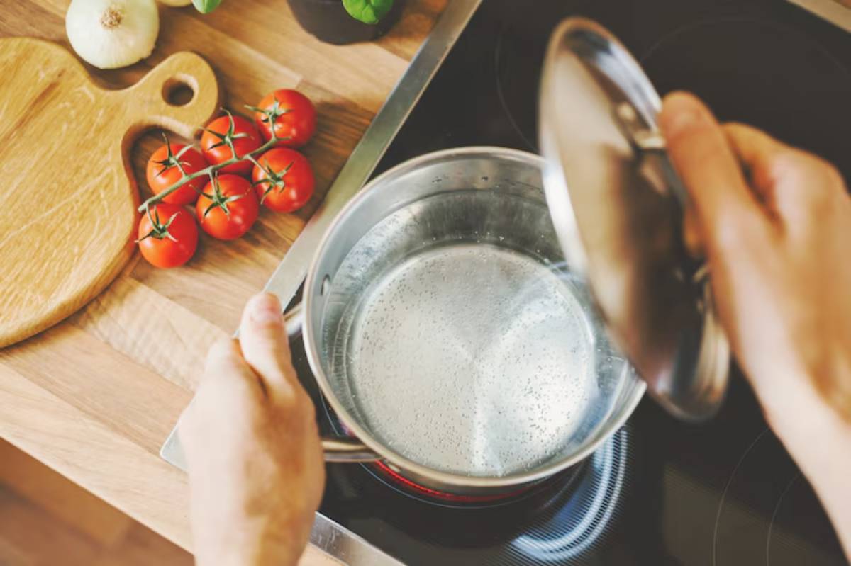 A person places a lid on a pot of boiling water, with fresh tomatoes and onions on a wooden cutting board nearby.