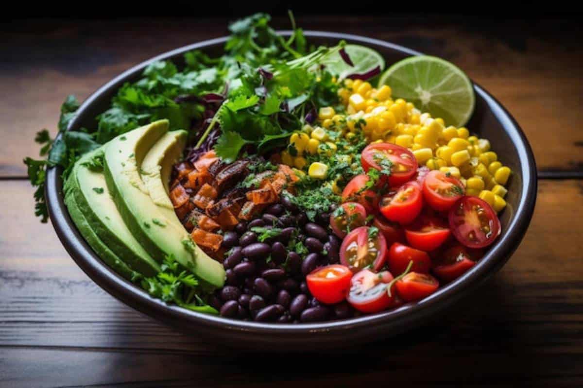 A vibrant salad bowl filled with avocado, black beans, corn, cherry tomatoes, and fresh herbs, garnished with lime wedges.