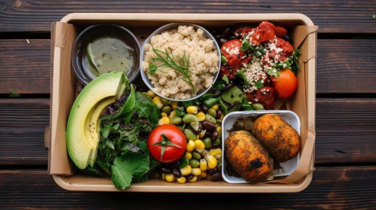 A vibrant lunch box featuring quinoa, salad greens, avocado, tomatoes, corn, black beans, and spiced potato balls with a green sauce.