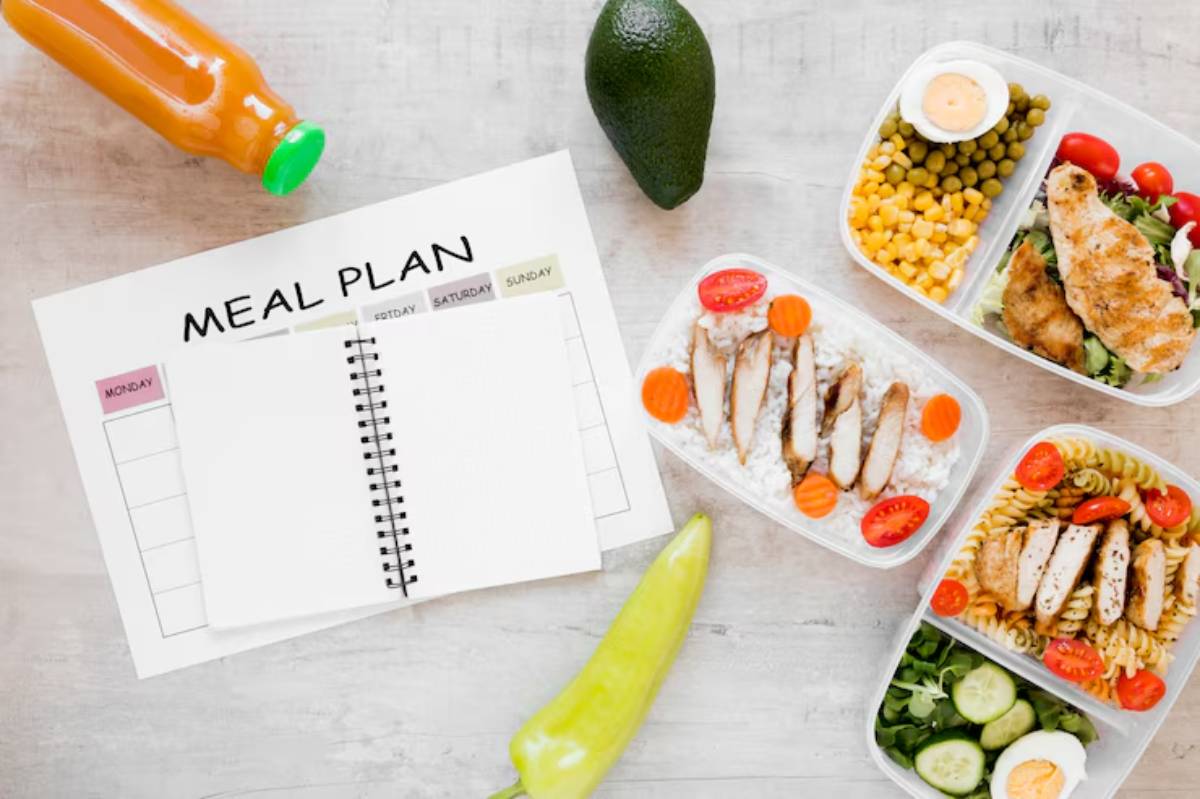 A meal planning setup featuring a blank planner, three balanced meal prep containers, an avocado, a pepper, and a juice bottle.