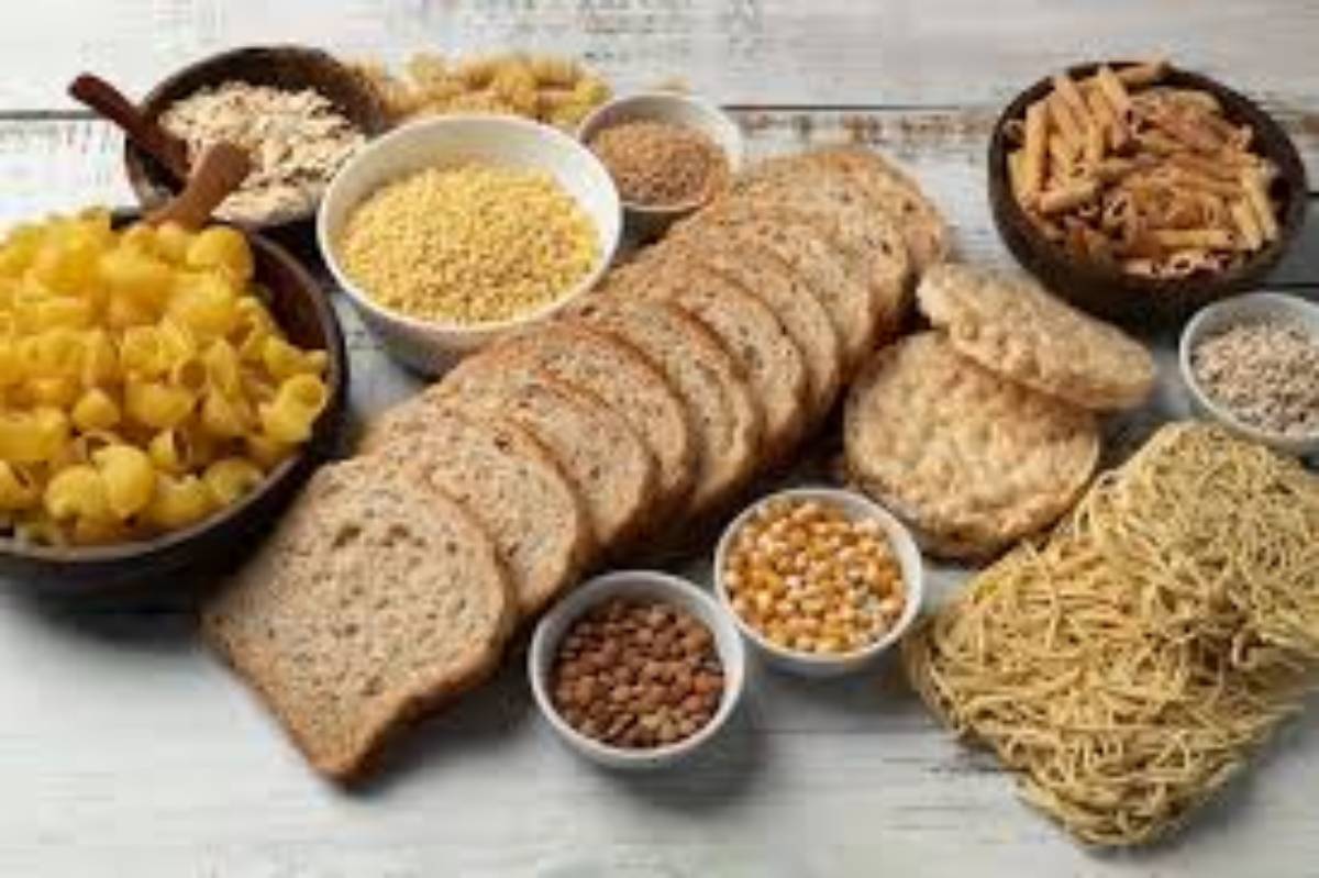 An assortment of grains and pasta, including sliced whole grain bread, macaroni, and various legumes in bowls on a rustic table.