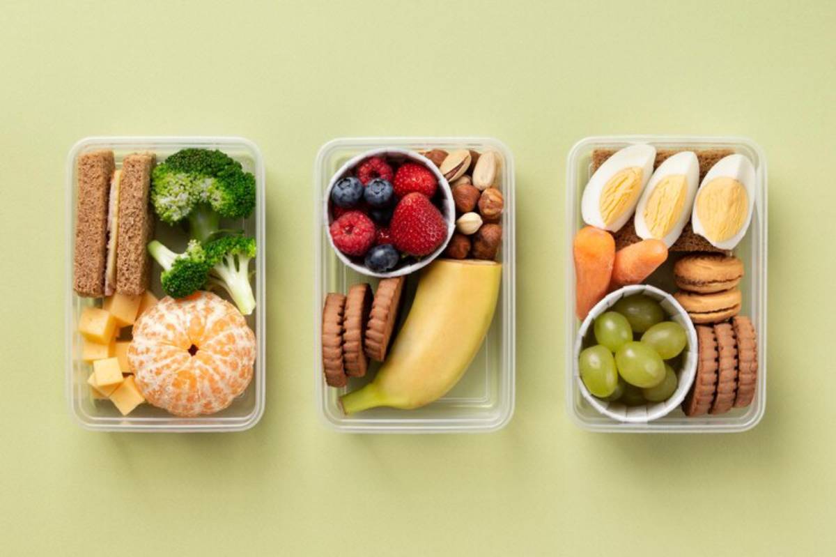 Three colorful lunch boxes filled with fruits, vegetables, nuts, cheese, boiled eggs, and cookies on a light green background.