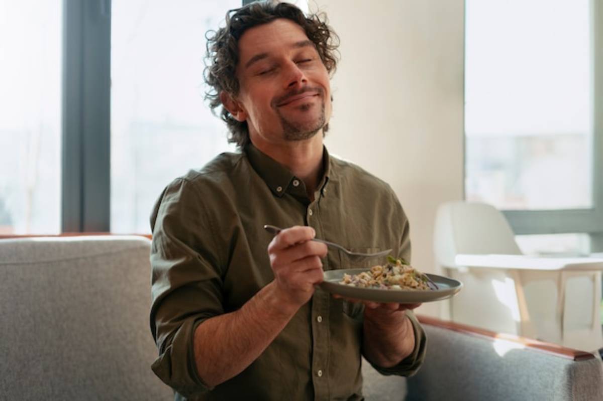 A person in a green shirt sits on a sofa, holding a plate of food with a fork, gazing thoughtfully out a large window.