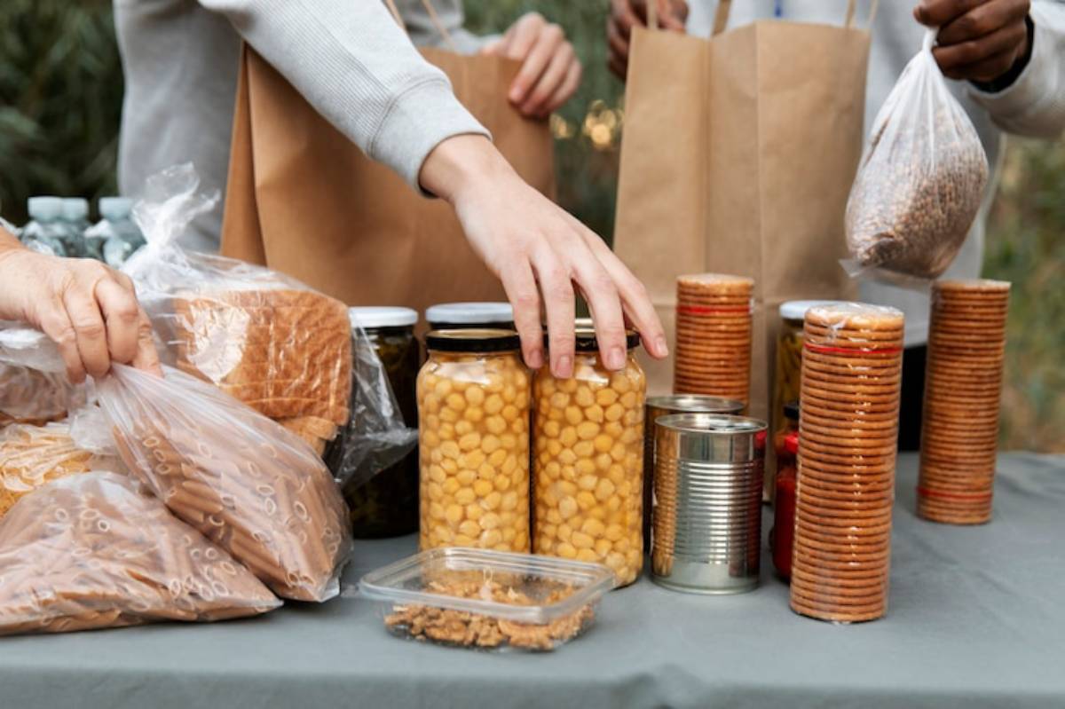 A person reaches for jars of food amid bags and cans of groceries on a table, displaying a variety of packaged pantry items.